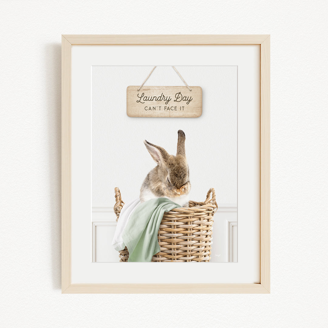 A framed image of a rabbit sitting in a wicker basket, with a wooden sign above it that says "Laundry Day Can't Face It".