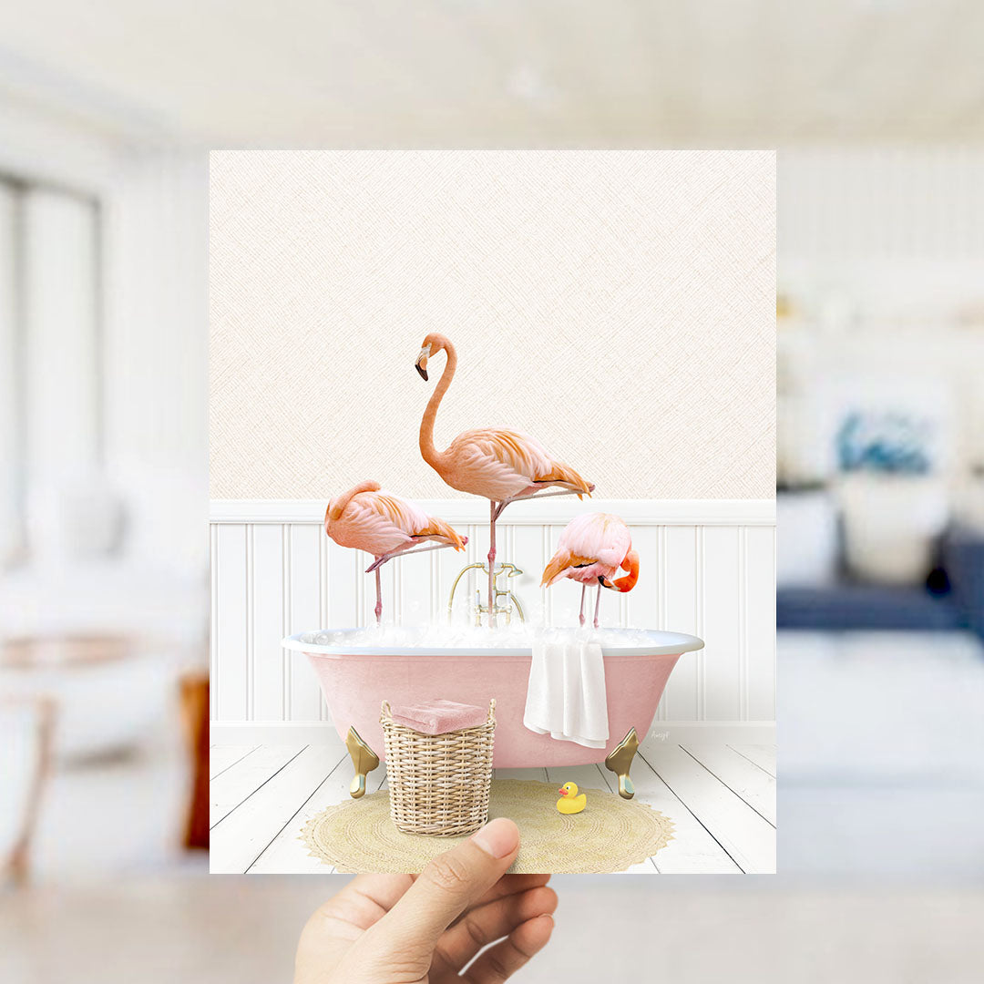 A hand holds up a framed image depicting three flamingos standing in a pink bathtub filled with bubbles, surrounded by a basket of bath toys and a yellow rubber duck.