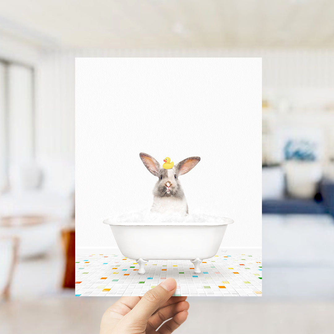 A hand holds up a framed image of a rabbit sitting in a bathtub filled with bubbles, wearing a yellow rubber duck.