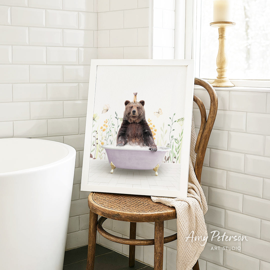 A framed print of a bear sitting in a bathtub is displayed on a wooden chair in a bathroom setting.