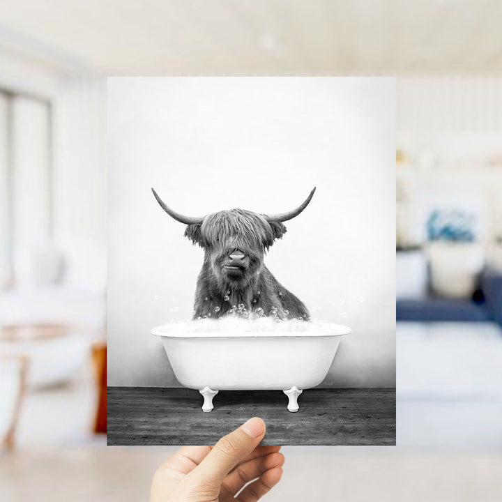 A black and white photograph of a Highland cow sitting in a bathtub filled with water.