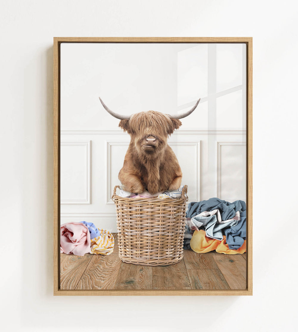 A brown Highland cow with shaggy hair sits in a wicker basket on a wooden floor, surrounded by various colorful clothes.