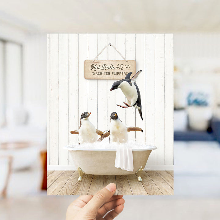 A hand holding a framed image of two penguins sitting in a bathtub, with a sign above them advertising hot baths for $2.00 and wash terriers.