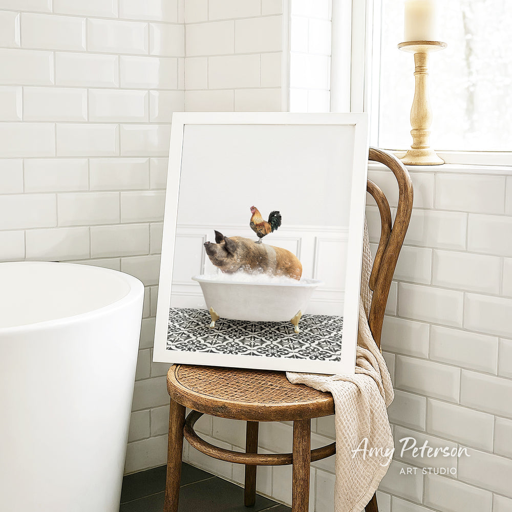 A framed image of a cat sitting in a bathtub, with a rooster perched on its head, is displayed on a wooden chair in a bathroom setting.