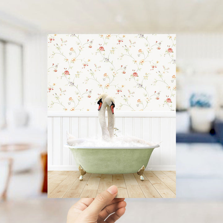 A hand holds up a framed photograph of a green bathtub filled with bubbles, with two swans sitting inside.