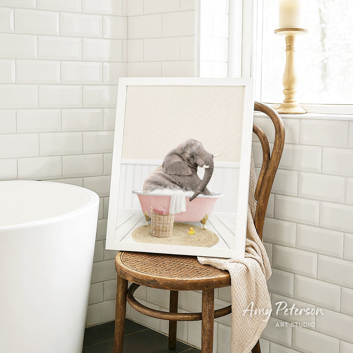 A framed image of a gray elephant sitting in a pink bathtub, surrounded by a basket of toys, is displayed on a wooden chair in a bathroom setting.