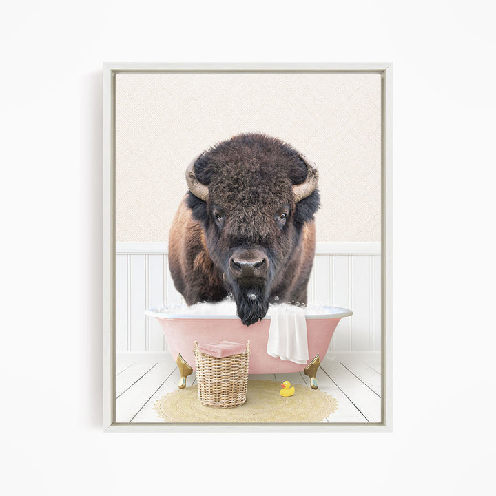 A buffalo with large horns is sitting in a pink bathtub filled with bubbles, surrounded by a yellow rubber duck and a basket.
