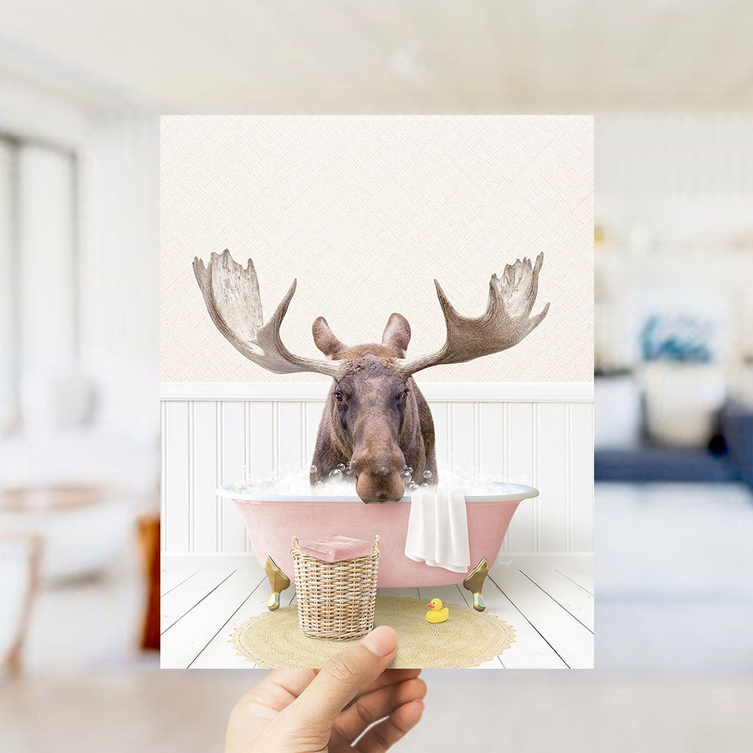 A moose with large antlers is sitting in a pink bathtub filled with water, surrounded by a yellow rubber duck and a wicker basket.
