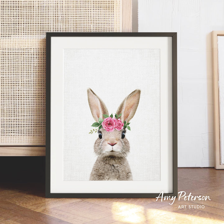 A framed print of a rabbit wearing a flower crown is displayed on a wooden floor against a white wall.