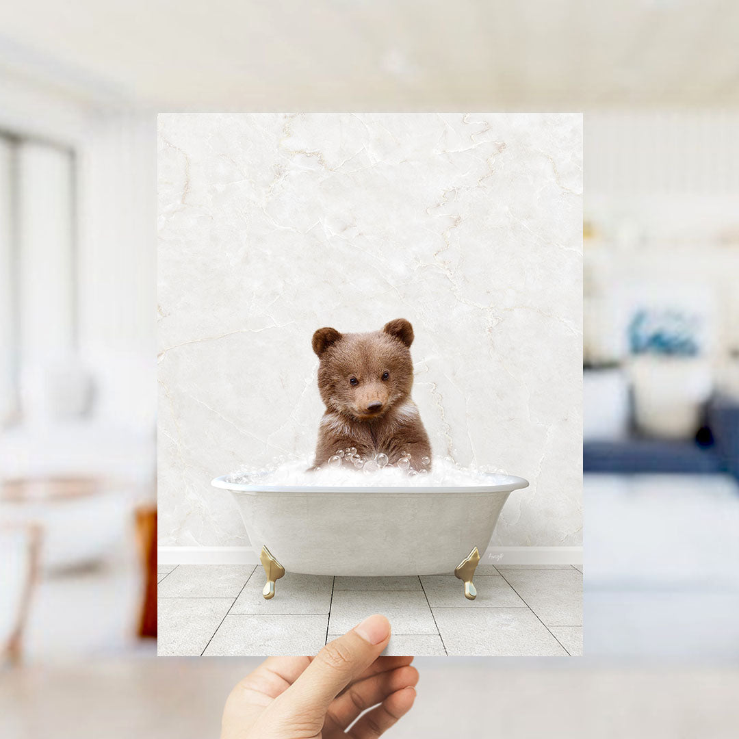 A hand holds up a framed image of a small brown bear sitting in a white bathtub filled with bubbles, creating a whimsical and playful scene.