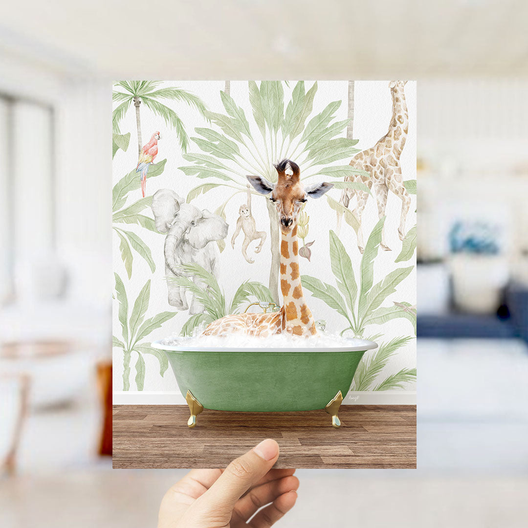A hand holds up a framed print featuring a giraffe sitting in a bathtub surrounded by tropical plants and animals, including a parrot, elephant, and monkey.