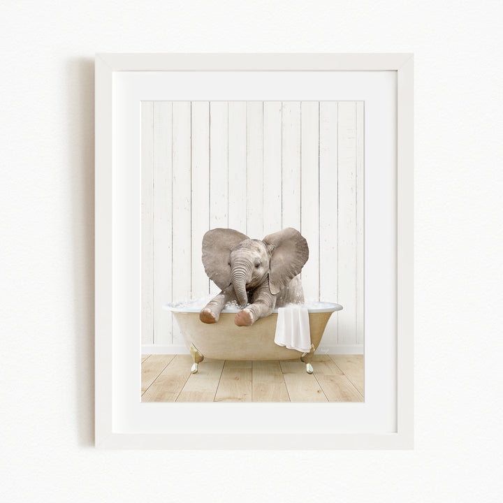 A baby elephant sitting in a bathtub, with its trunk curled up and ears spread out, against a white wooden wall.