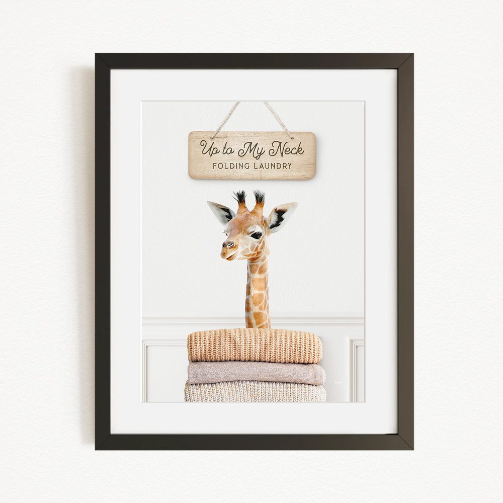 A framed image of a giraffe standing on a pile of folded clothes, with a sign above that says "Up to My Neck Folding Laundry".