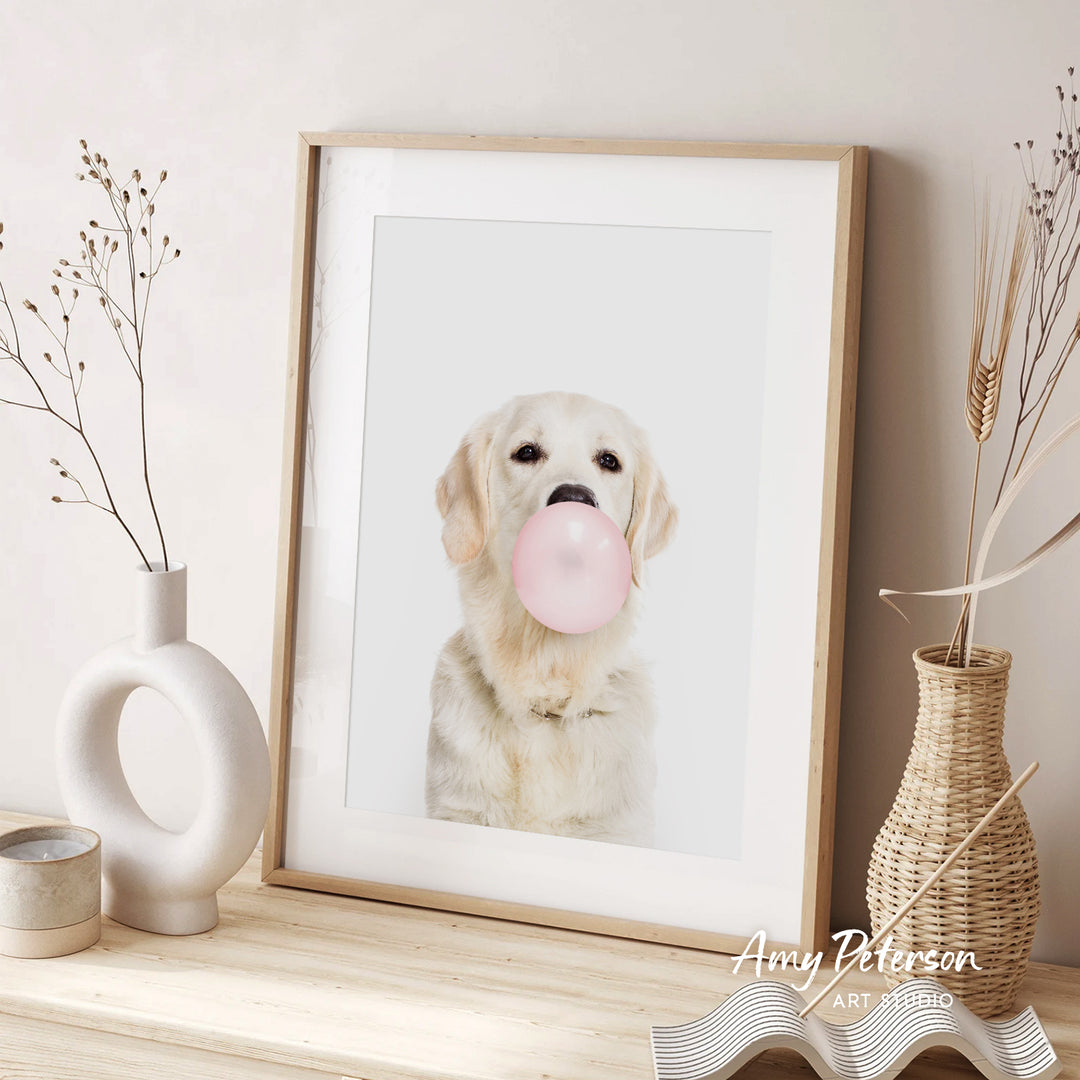 A framed photograph of a golden retriever dog blowing a bubble with a pink bubble gum bubble, accompanied by decorative elements such as a white vase and a woven basket.