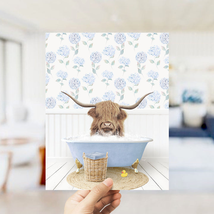 A hand holds up a framed image depicting a Highland cow with long horns sitting in a blue bathtub filled with bubbles, surrounded by a floral patterned wall.