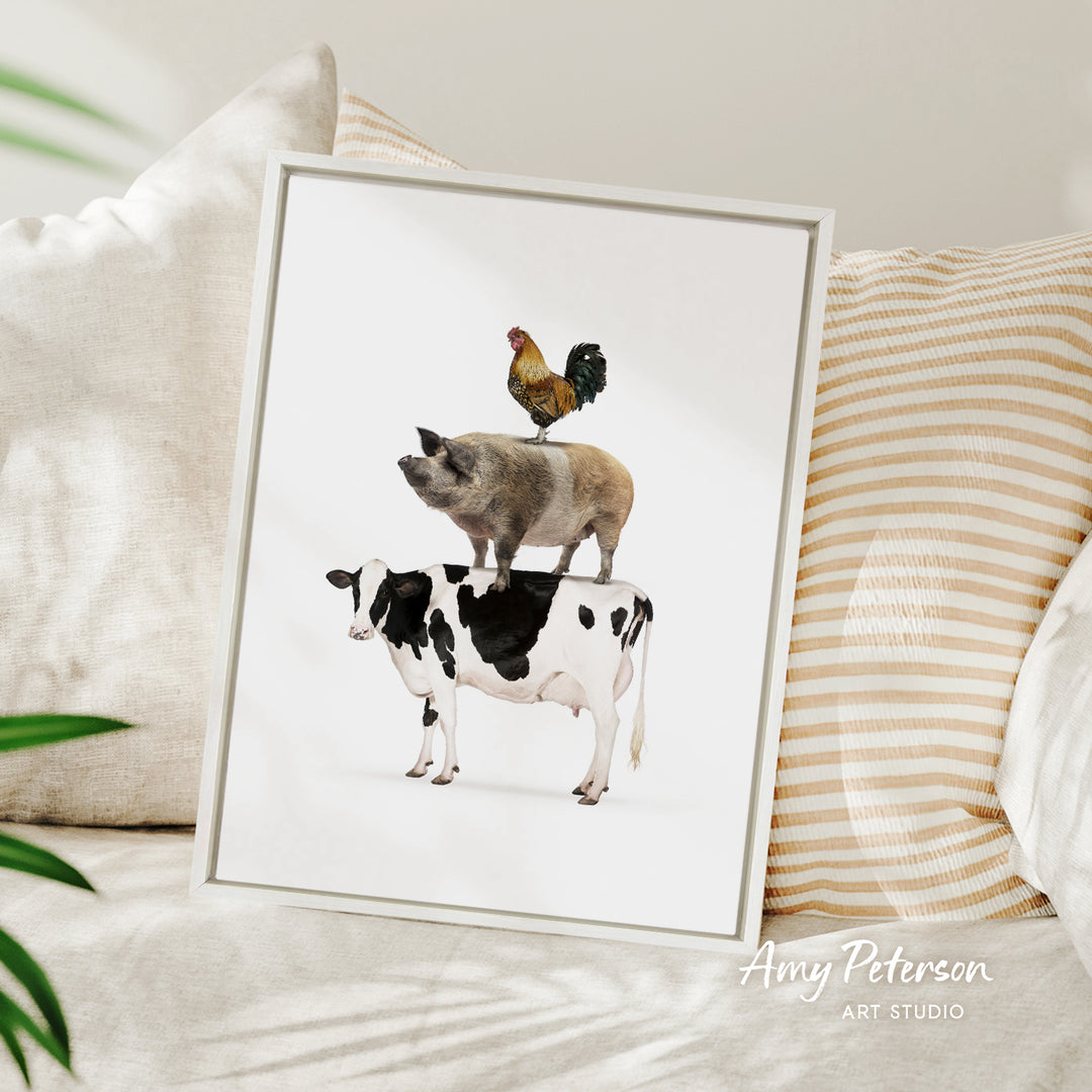 A framed illustration of a pig, cow, and rooster standing on top of each other, with a white background and a striped pillow in the background.