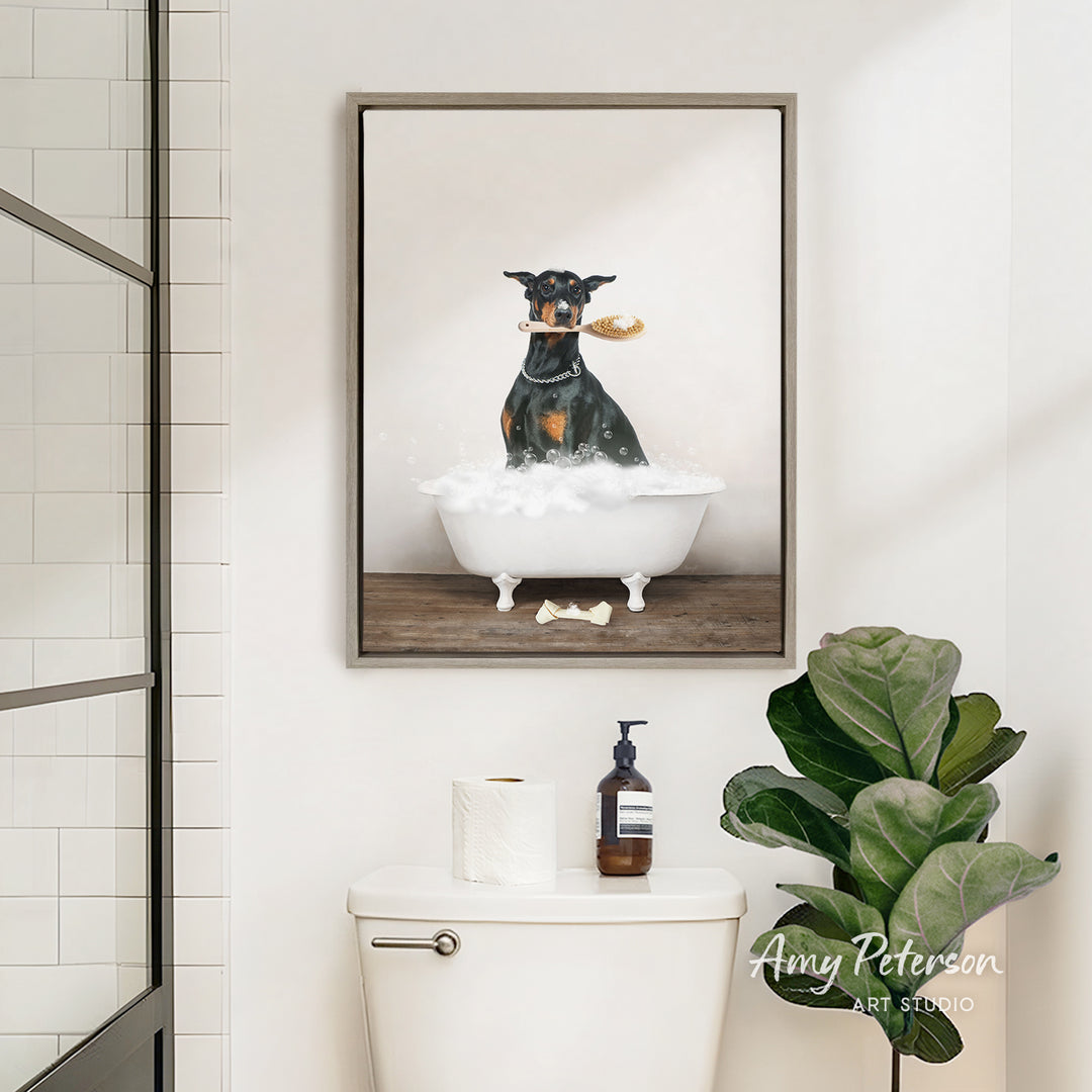 A framed image of a dog sitting in a bathtub, holding a toy in its mouth, is hanging on a bathroom wall above a toilet.