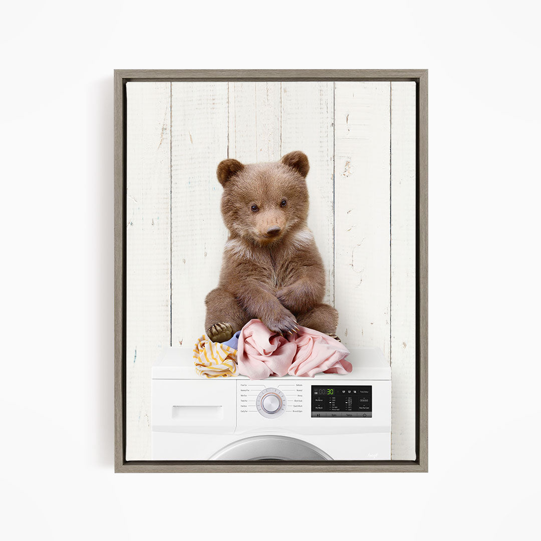 A brown bear sitting on top of a white washing machine, with clothes and a toy nearby.