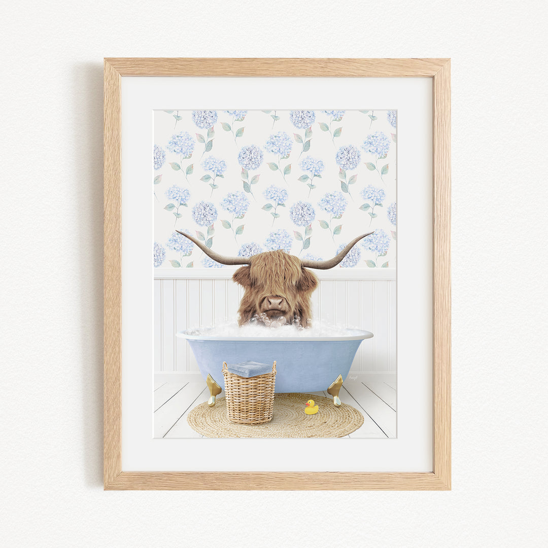 A framed image depicting a Highland cow with long horns sitting in a blue bathtub filled with water, surrounded by a floral patterned wall.