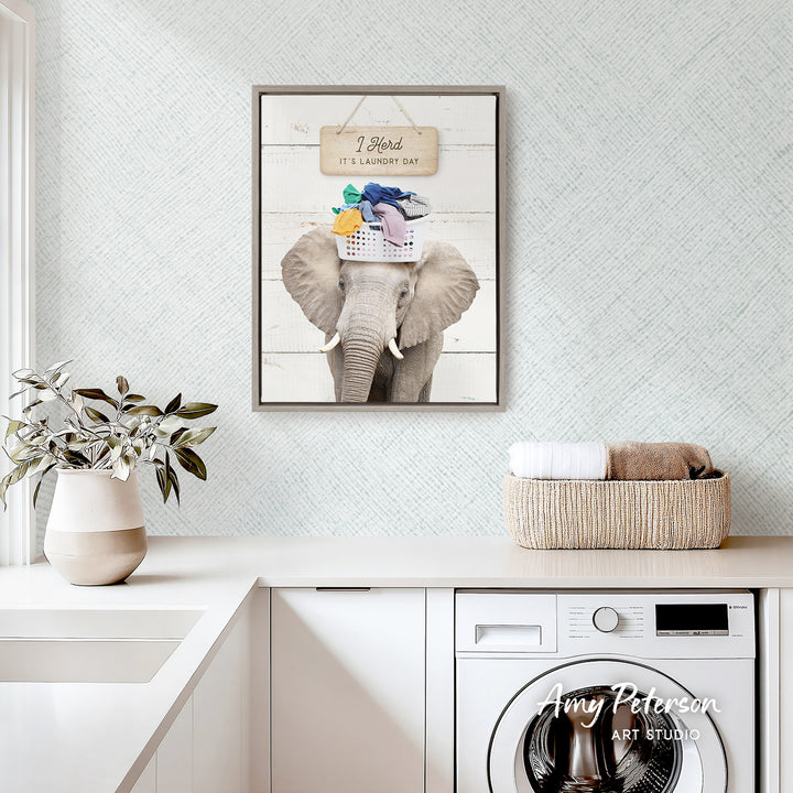a framed picture of an elephant with a basket of laundry on its head, hanging on a wall above a white countertop with a potted plant and a basket of towels.