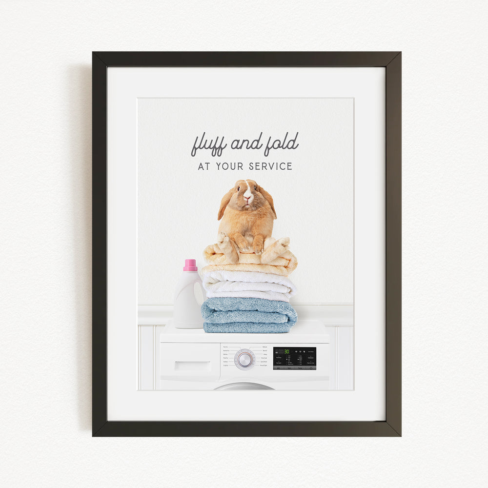 A framed image depicting a fluffy rabbit sitting atop a stack of folded towels, with a bottle of laundry detergent nearby.