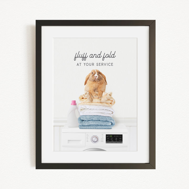 A framed image depicting a fluffy rabbit sitting atop a stack of folded towels, with a bottle of laundry detergent nearby.