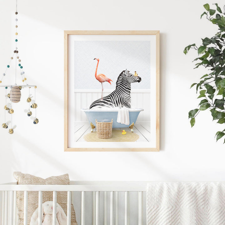 A framed illustration of a zebra sitting in a bathtub, with a pink flamingo standing on its back, is displayed on a white wall in a nursery setting.