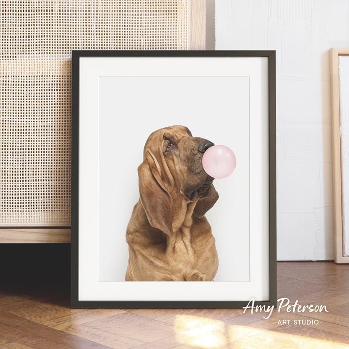 A brown dog is blowing a bubble with a pink bubble gum bubble in a framed photograph.