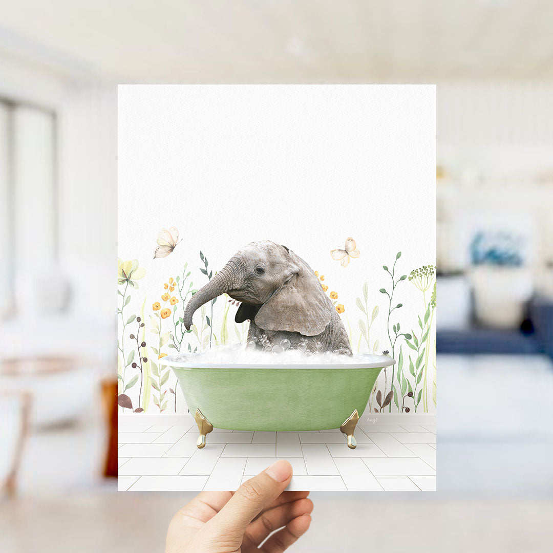 A hand holds up a framed image depicting a baby elephant sitting in a green bathtub filled with bubbles, surrounded by flowers and butterflies.