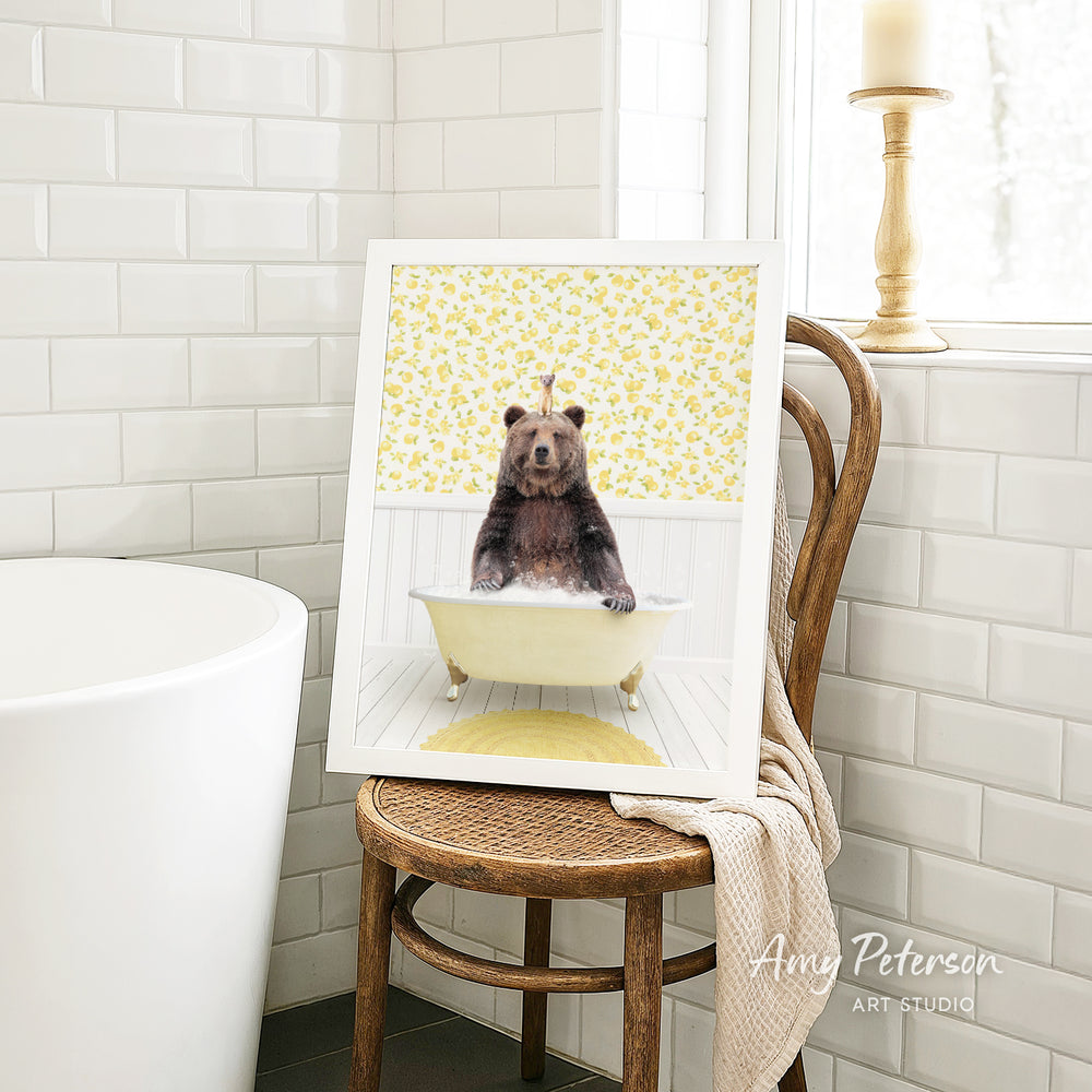 A framed image of a bear sitting in a bathtub, surrounded by yellow flowers, is displayed on a wooden chair in a bathroom setting.