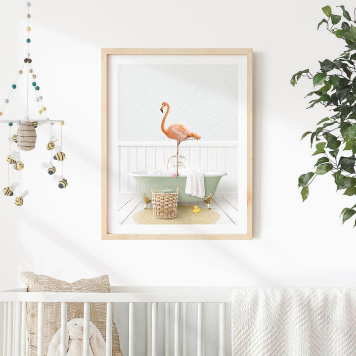 A framed print of a pink flamingo standing in a bathtub is displayed on a white wall, with a white crib and a hanging mobile in the background.