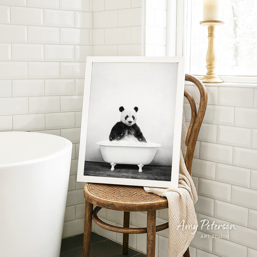 A framed black and white photograph of a panda sitting in a bathtub is displayed on a wooden chair in a bathroom setting.
