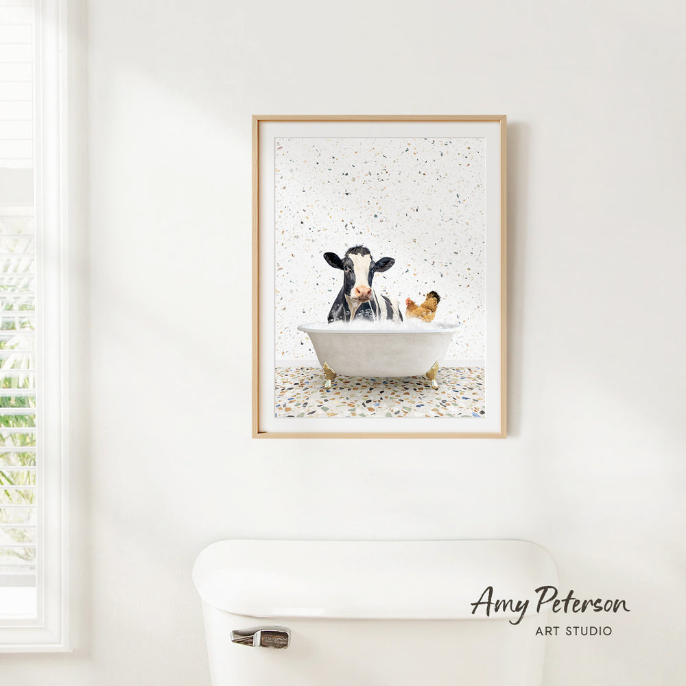 A framed print of a cow sitting in a bathtub, with a chicken in the tub, is hanging on a white wall.