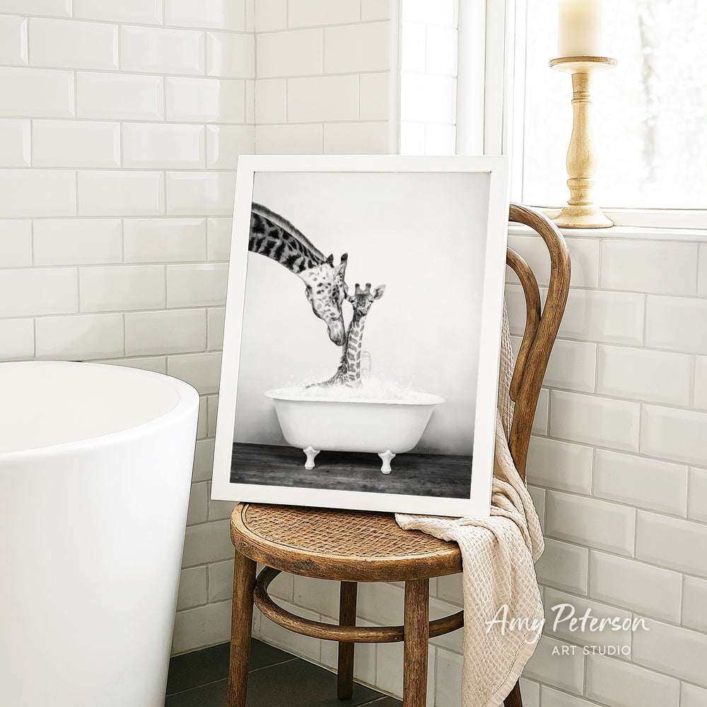 A framed black and white photograph of a giraffe standing in a bathtub, with another giraffe partially submerged in the tub, is displayed on a wooden chair in a bathroom setting.