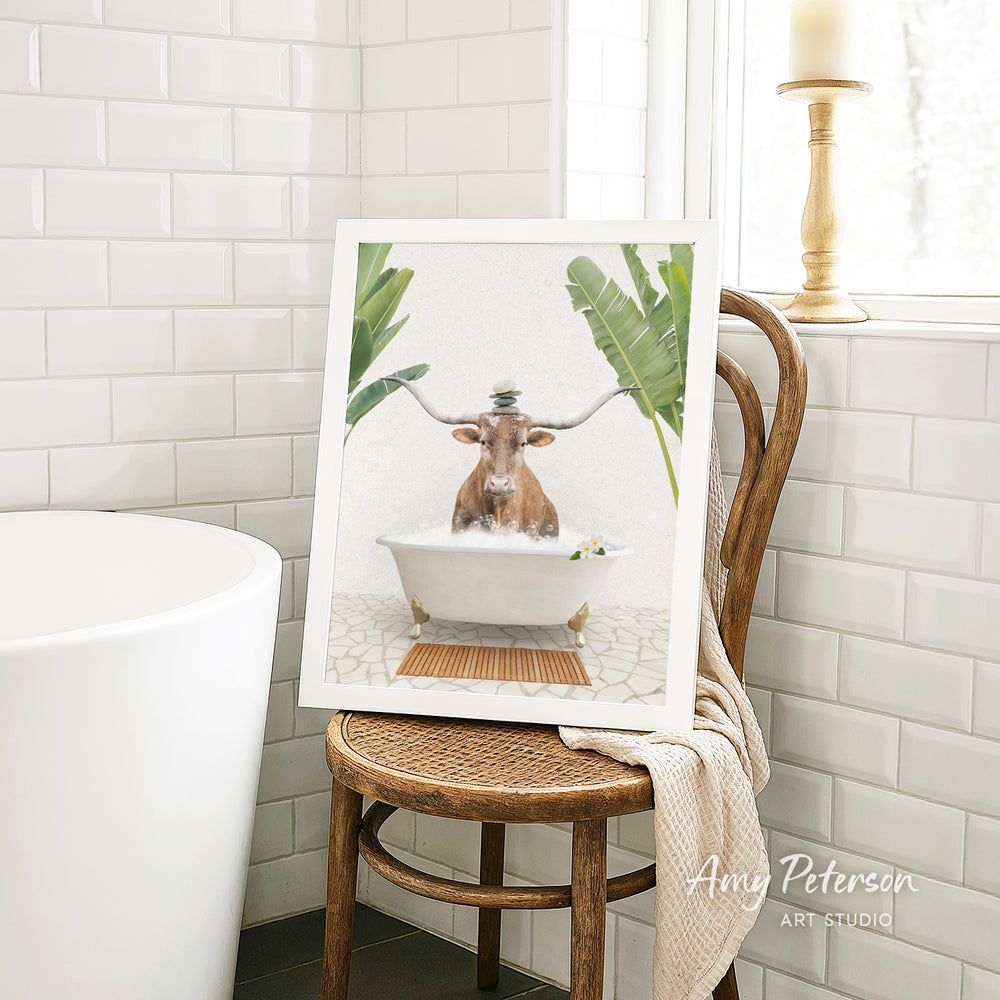A framed image of a cow sitting in a bathtub, surrounded by tropical plants, is displayed on a wooden chair in a bathroom setting.