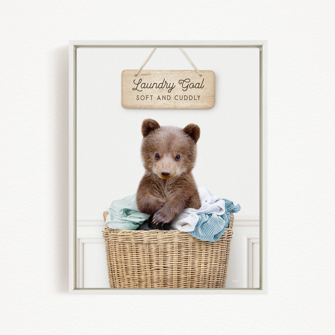 A cute brown bear sitting in a wicker laundry basket, with a wooden sign above that says "Laundry Goal Soft and Cuddly".