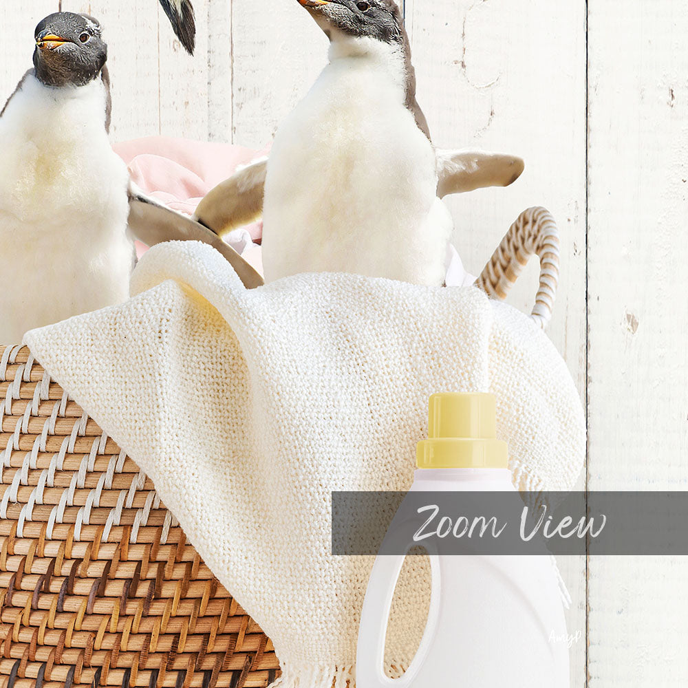 A woven basket containing two penguins, with a white cloth and a yellow bottle of detergent in the foreground.