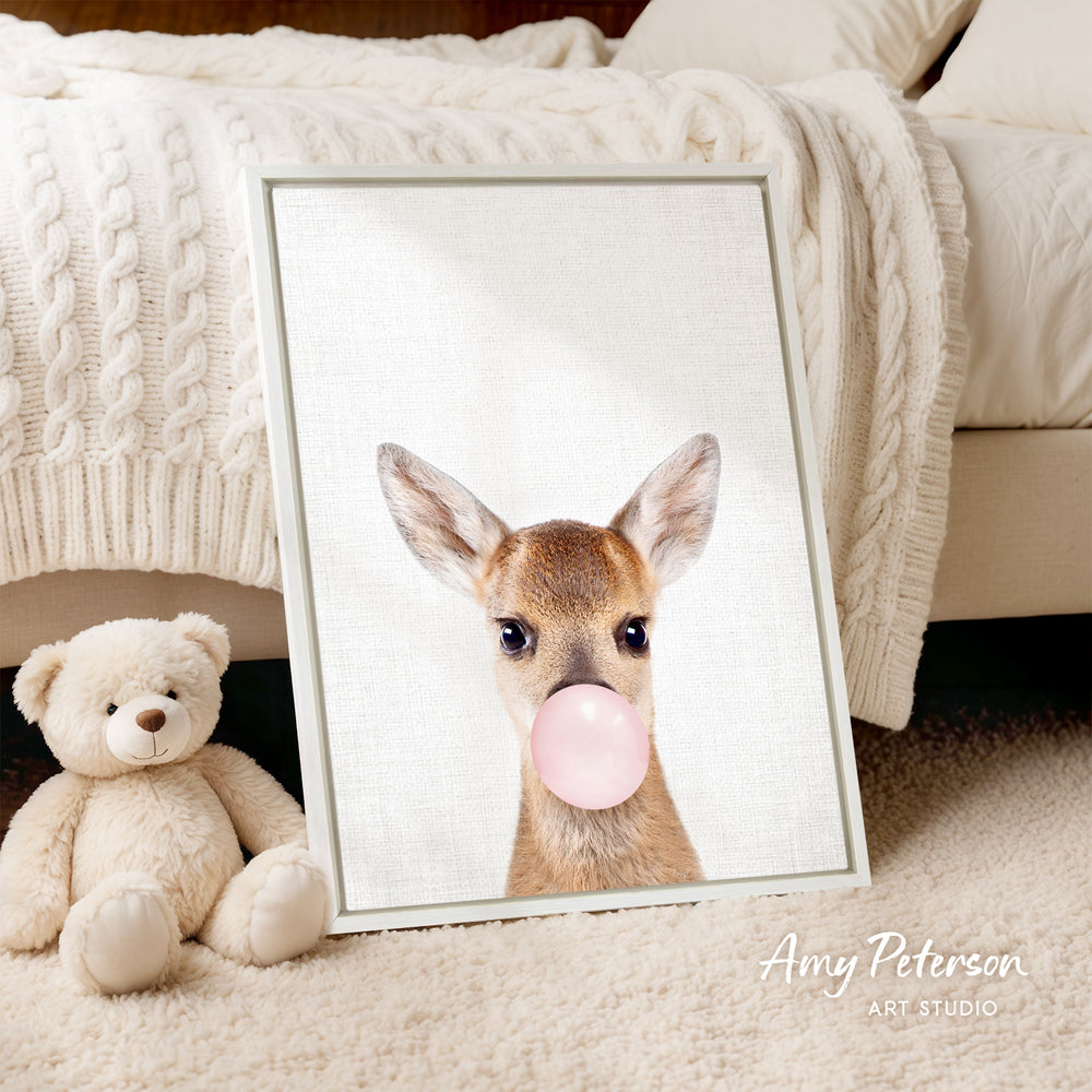 A framed print of a deer blowing a bubble gum bubble is displayed on a bed, accompanied by a teddy bear.