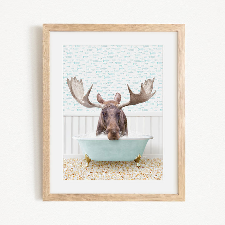 A moose with large antlers is sitting in a light blue bathtub, surrounded by a patterned tile floor and a white wall with a blue and white fish wallpaper.