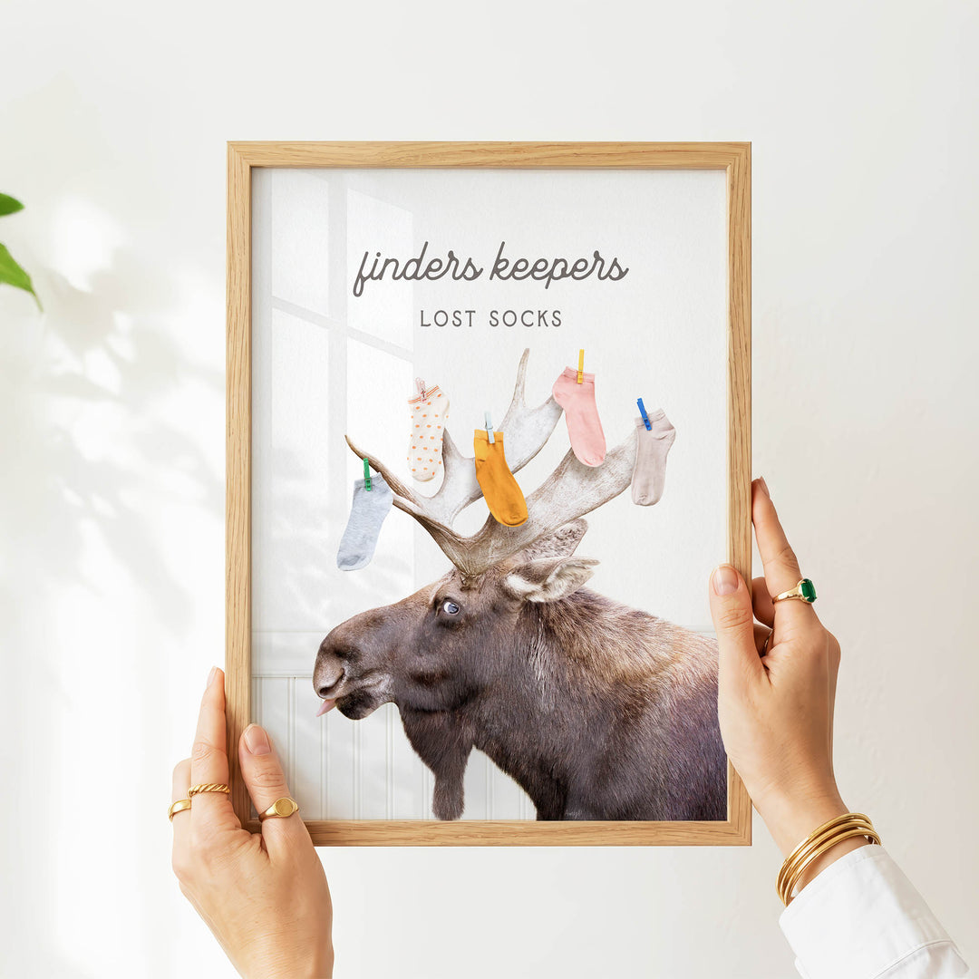 A person's hands are holding a framed picture of a moose with a sign that says "finders keepers lost socks".