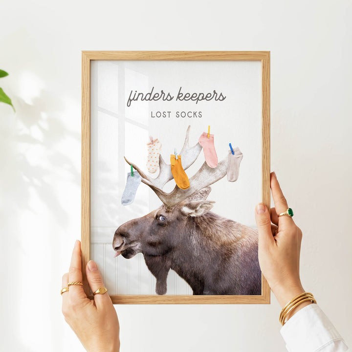 A person's hands are holding a framed picture of a moose with a sign that says "finders keepers lost socks".