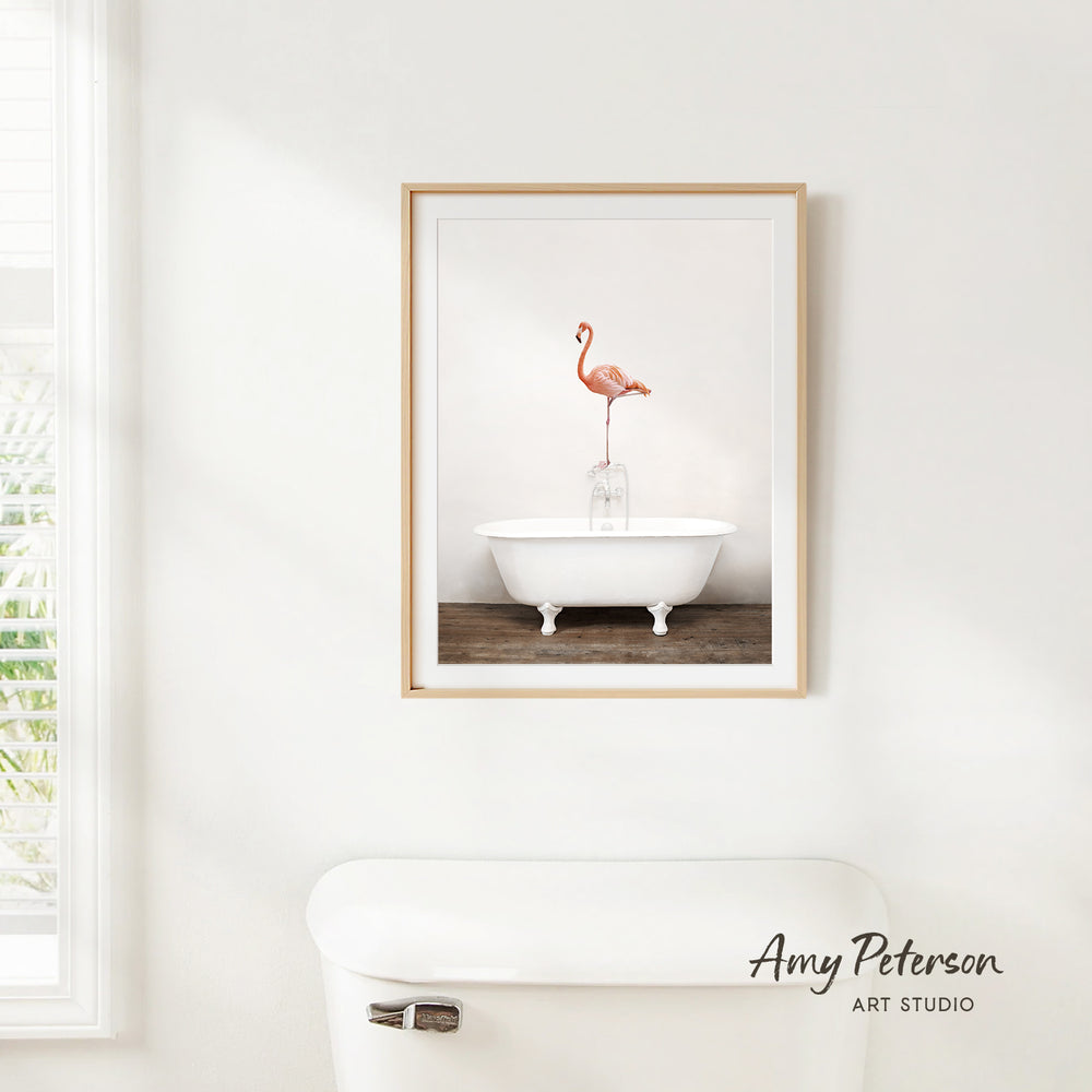 A framed image of a pink flamingo standing on a white bathtub is hanging on a white wall in a bathroom.