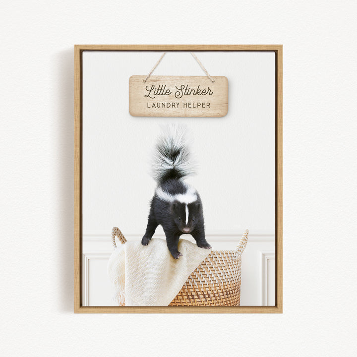 A framed image of a black and white skunk standing on a woven basket, with a wooden sign above it that reads "Little Stinker Laundry Helper".