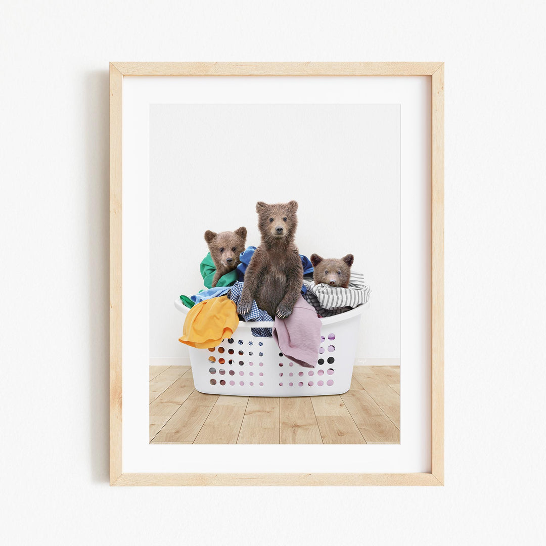 A framed image depicting three brown bears sitting in a laundry basket filled with clothes.