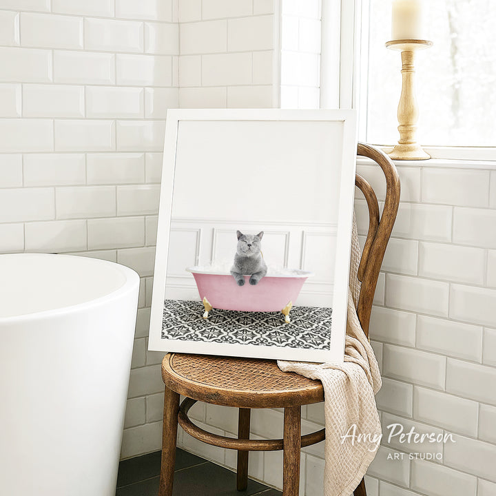 A framed image of a gray cat sitting in a pink bathtub, with a wooden chair and a white tiled wall in the background.