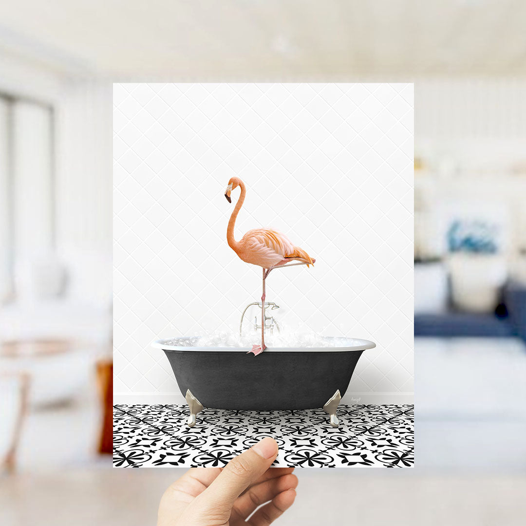 A hand holds up a framed image of a pink flamingo standing on a black bathtub filled with water, set against a white background with a black and white patterned floor.