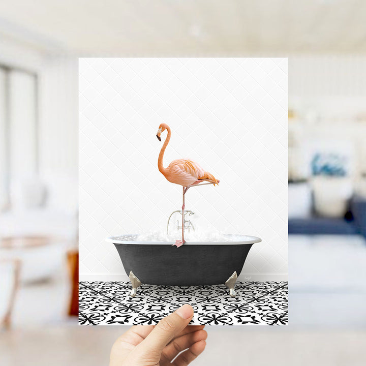 A hand holds up a framed image of a pink flamingo standing on a black bathtub filled with water, set against a white background with a black and white patterned floor.