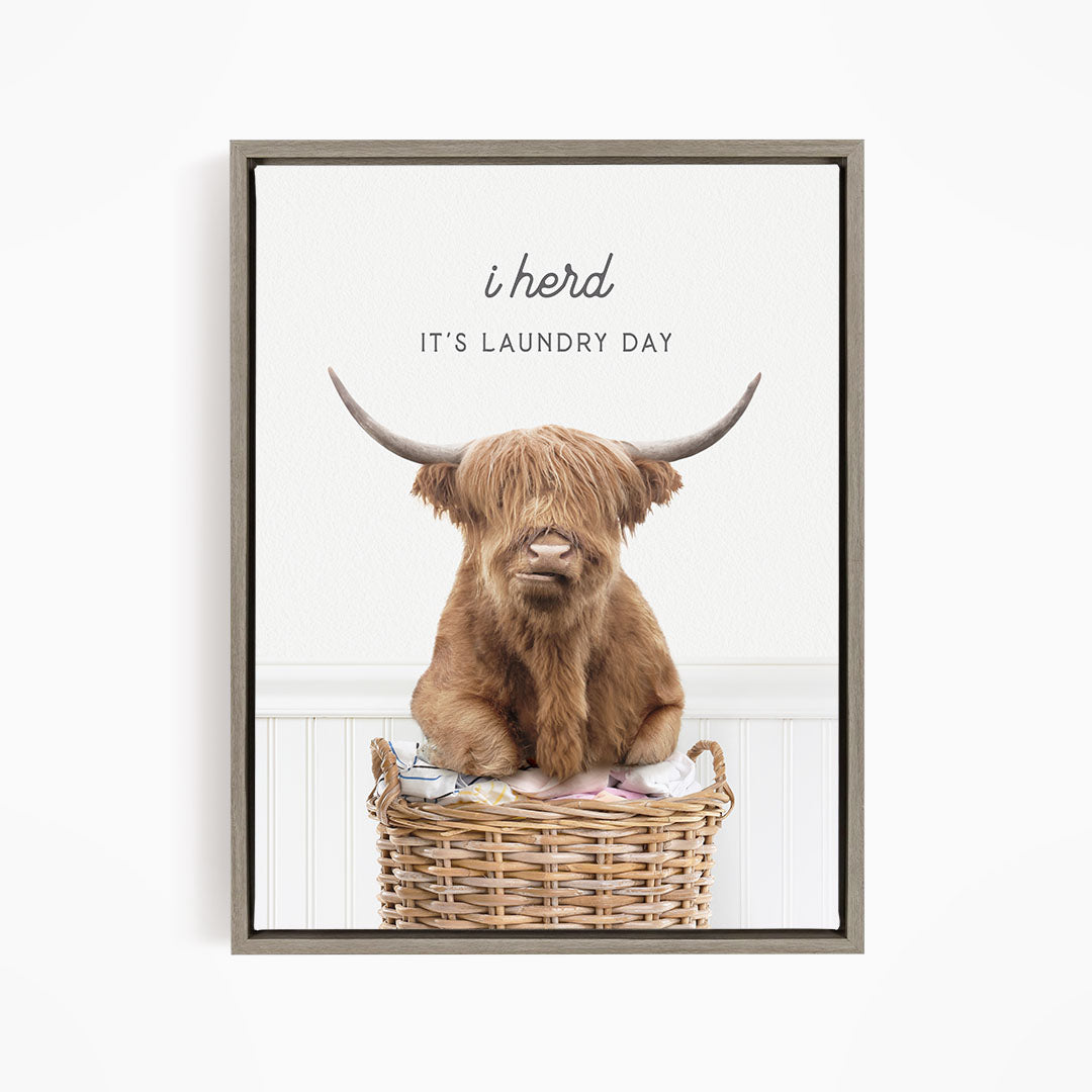 A brown Highland cow with large horns is sitting in a wicker basket, with the text "It's Laundry Day" above the image.