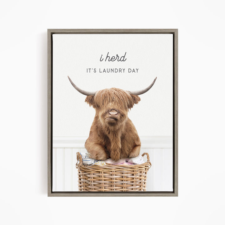A brown Highland cow with large horns is sitting in a wicker basket, with the text "It's Laundry Day" above the image.