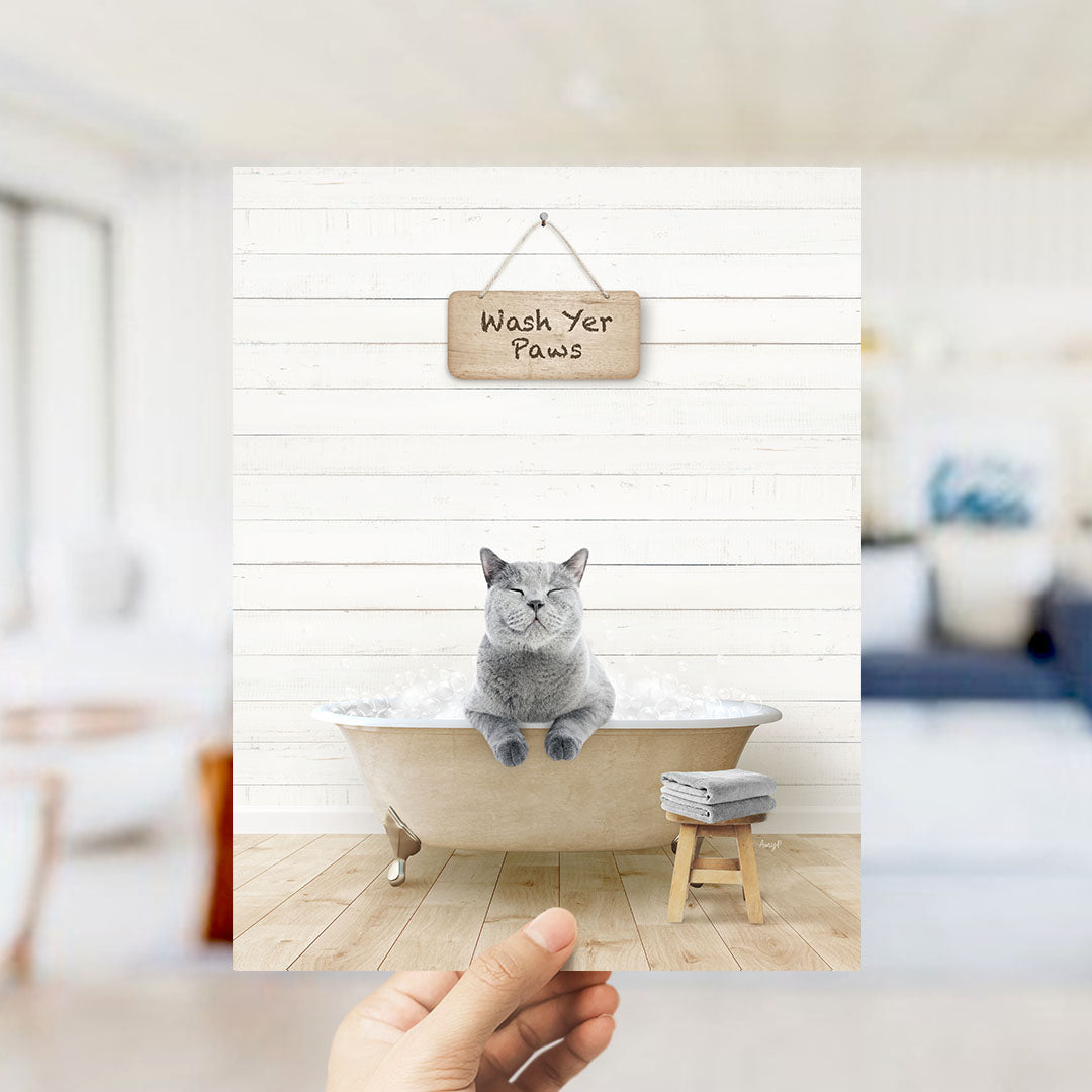 A gray cat sitting in a bathtub filled with bubbles, with a wooden sign above that says "Wash Yer Paws".